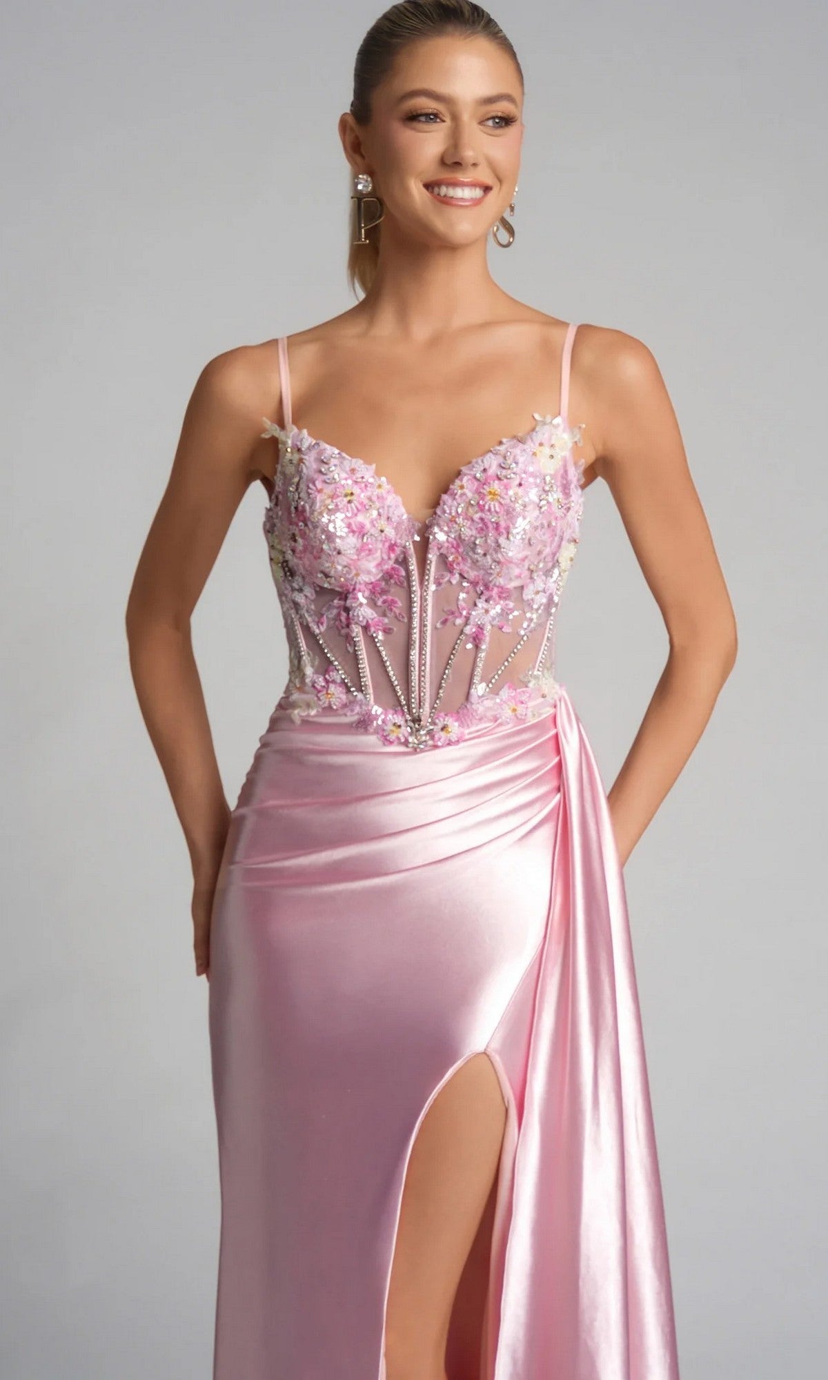 A woman models the Portia and Scarlett PS26270 long prom dress, featuring light pink satin, a floral applique bodice, spaghetti straps, and a thigh-high slit against a plain background.